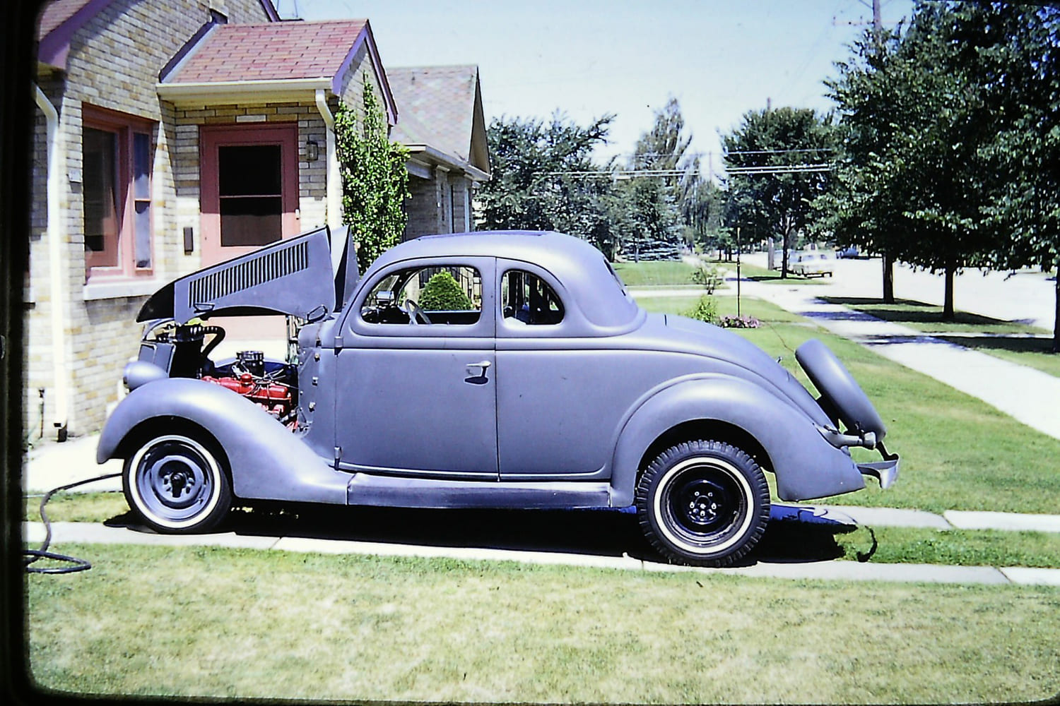 Historic color slide showing the driver-side profile of a primer-gray 1936 Ford five-window coupe with open hood parked on a suburban lawn in 1967.