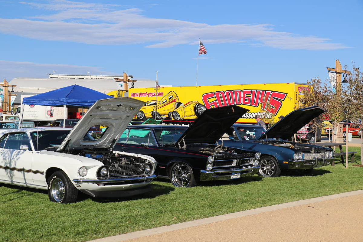 A white 1969 Mustang, black Pontiac GTO, and dark blue muscle car parked on grass with their hoods open in front of the yellow Goodguys hauler.