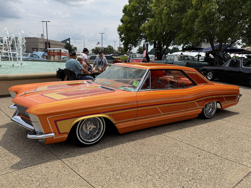 Low front three-quarter view of a bagged sunset orange 1965 Buick Riviera featuring intricate multi-color pinstriping, custom panel paint, and wire wheels by a fountain.