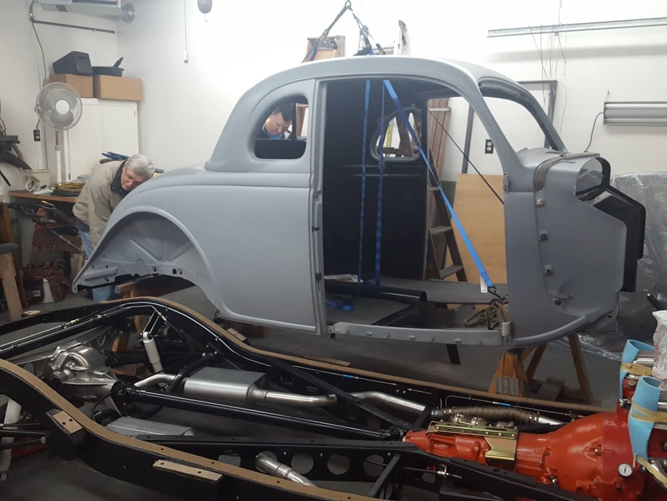 Restoration workshop scene showing a primer-gray 1936 Ford coupe body suspended on lift straps directly above a fully assembled black rolling chassis.