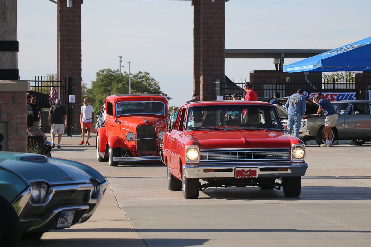 Front view of a bright red 1966 Chevy II Nova sedan cruising down a concrete walkway ahead of an orange street rod coupe