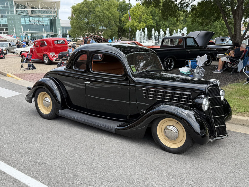 Side profile of a lowered high-gloss black 1936 Ford five-window coupe with tan artillery-style wheels parked on asphalt outside the exhibition center.