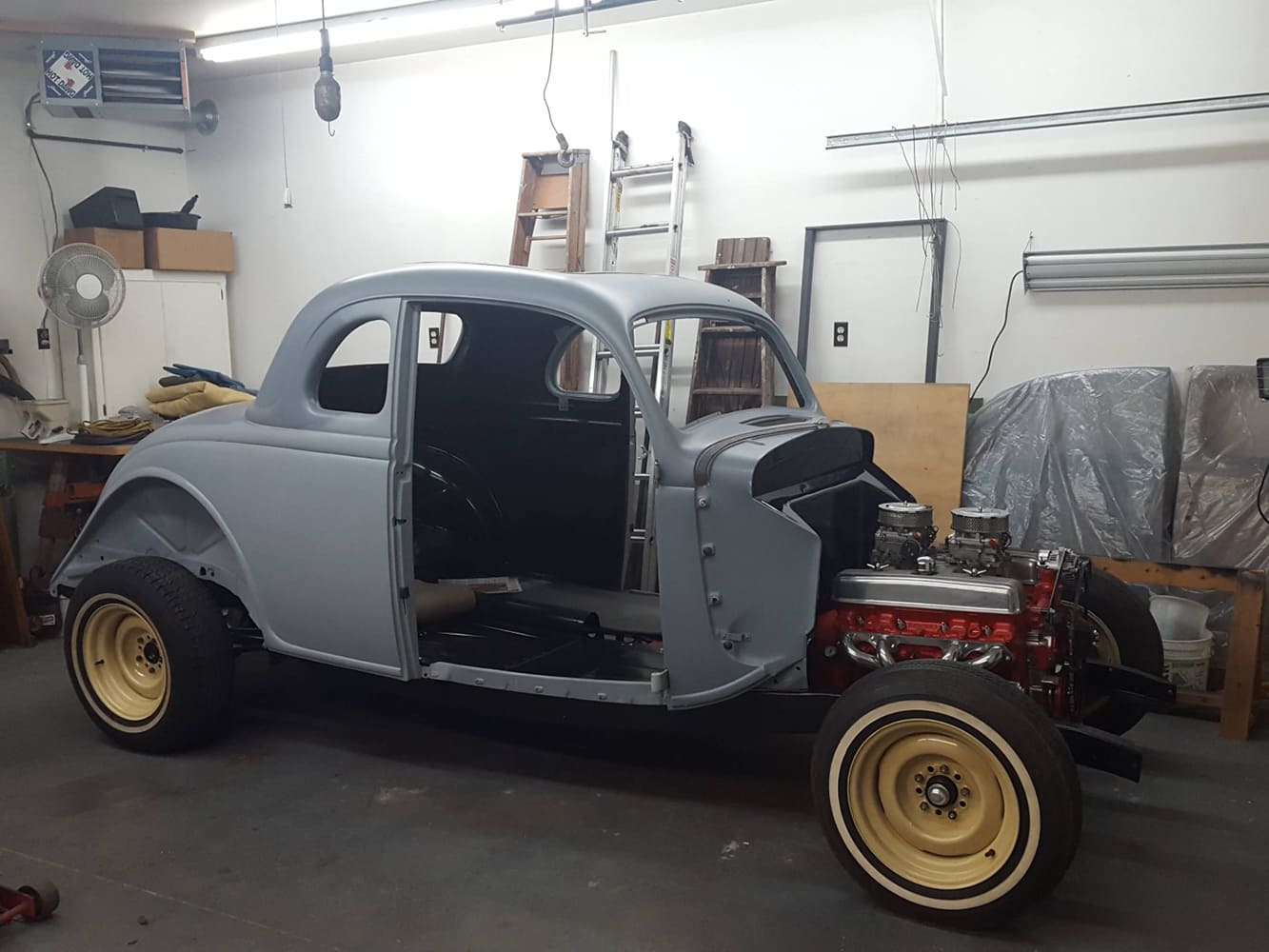 Three-quarter side view of a primer-gray 1936 Ford coupe shell bolted to its rolling frame with cream wheels and an exposed orange V8 engine.