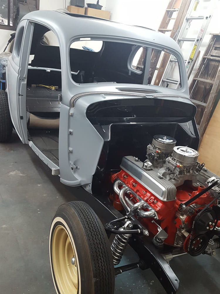 Front three-quarter angle of a 1936 Ford coupe chassis build displaying an orange dual-quad engine, tubular headers, independent front suspension, and cream artillery wheel.