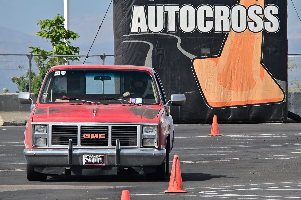 Front view of a red squarebody GMC pickup truck navigating orange traffic cones on an autocross course in front of an inflatable banner.