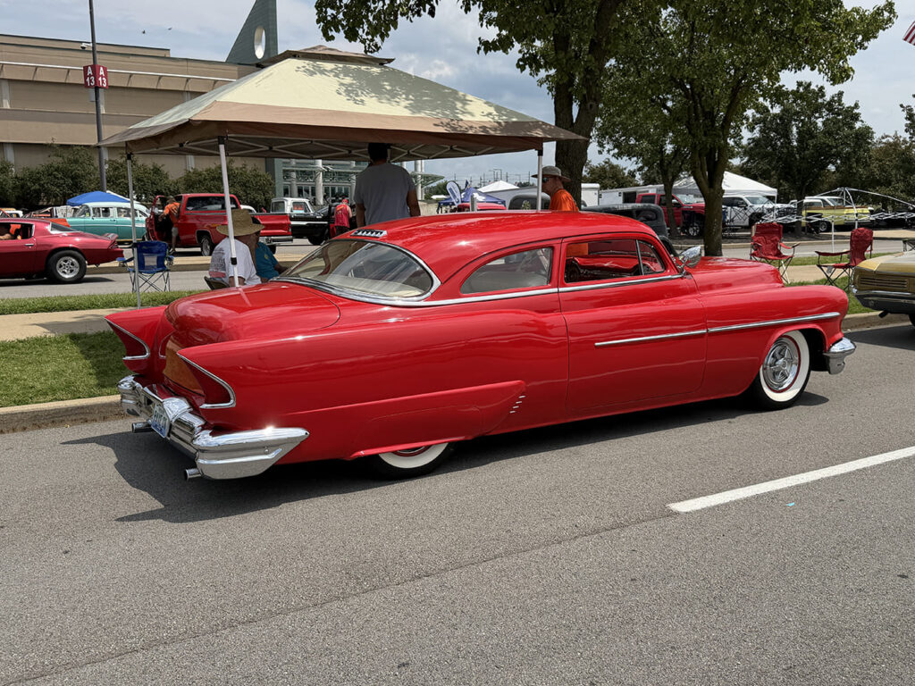 Rear three-quarter perspective of a bright red 1954 Oldsmobile Holiday coupe with chrome fender skirts, shaved body lines, and wide whitewall tires.