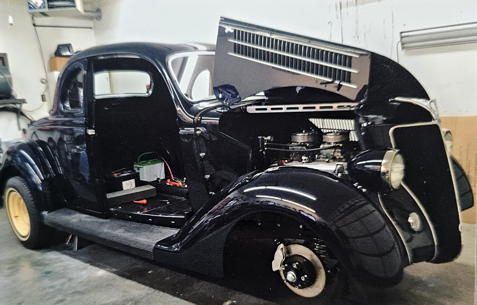 Front three-quarter view of a freshly painted dark blue 1936 Ford coupe undergoing final assembly with open hood sides in a home garage.