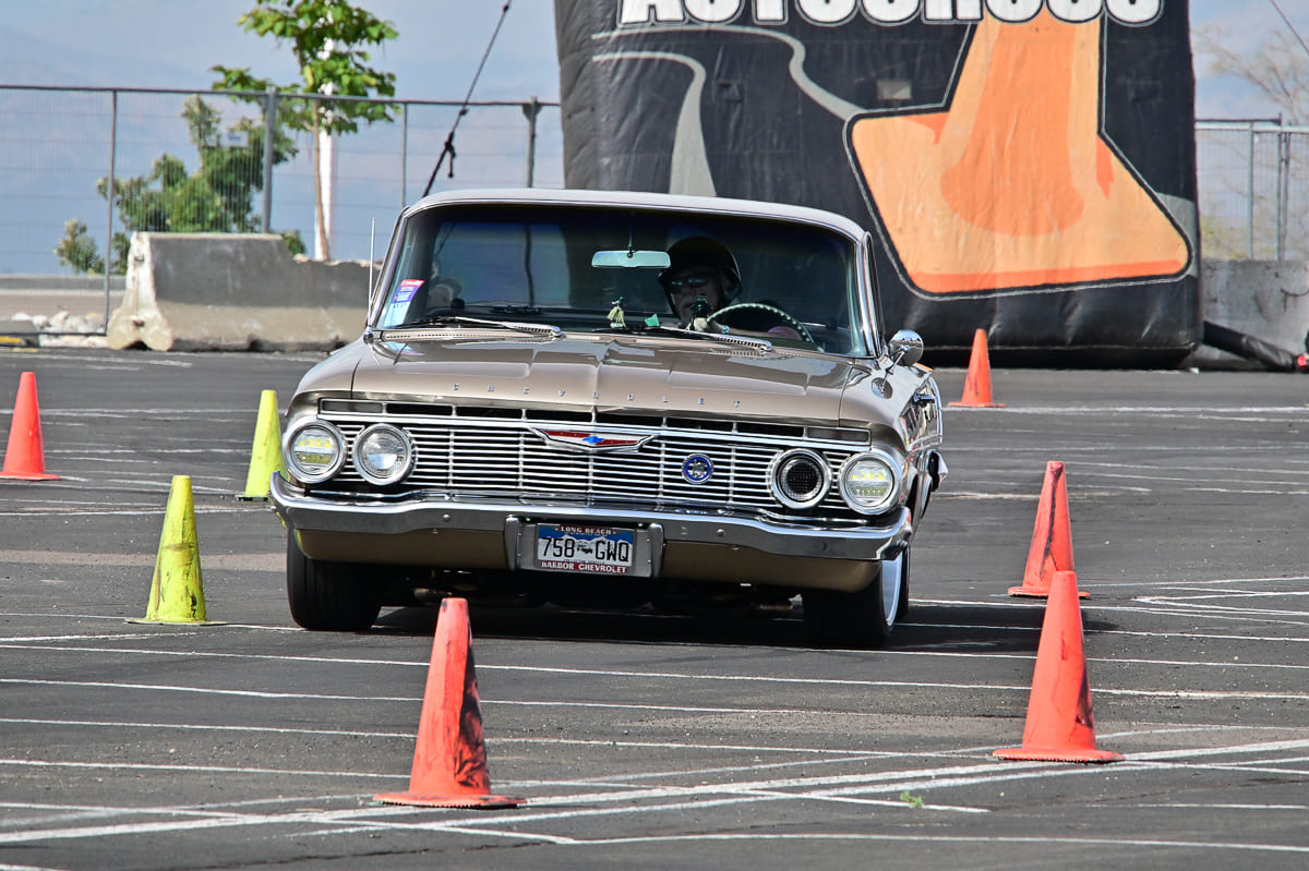 Bronze-gold 1961 Chevrolet sedan maneuvering tightly through cone gates on an asphalt autocross track.