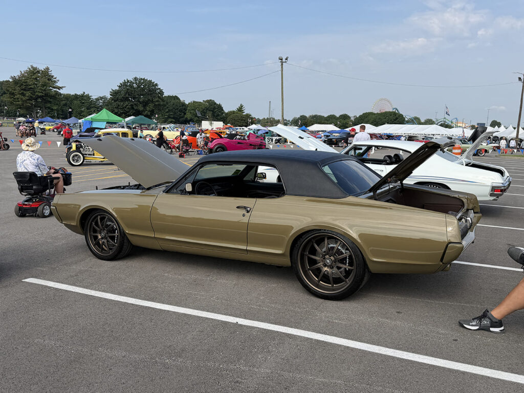 High rear three-quarter view of a pro-touring metallic gold Mercury Cougar with a black vinyl roof, open hood and trunk, and bronze performance wheels.