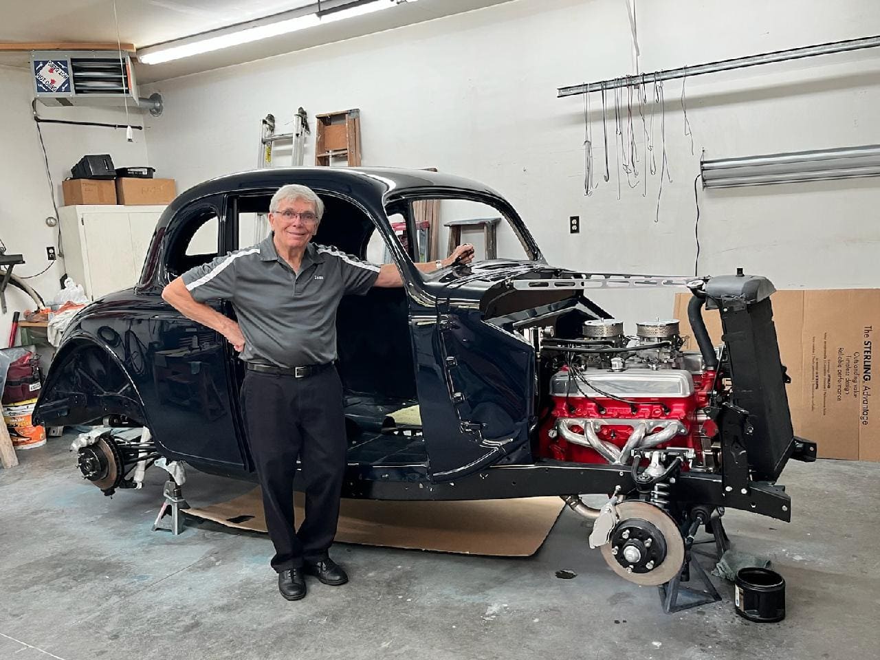 Tom Lawrence smiling beside his midnight blue 1936 Ford coupe project resting on jack stands with exposed dual-carb engine and front disc brakes.