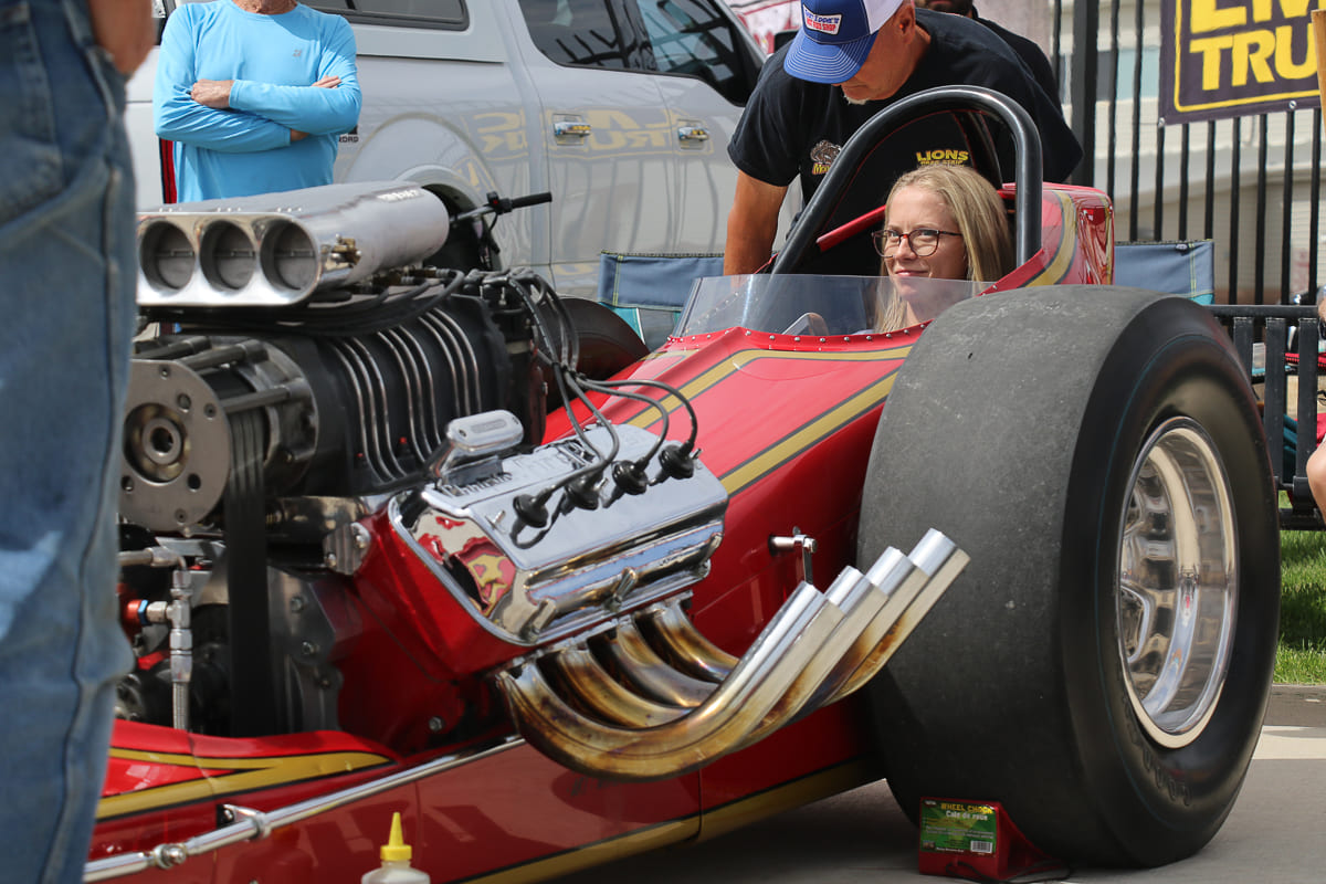 Close-up of a female driver seated behind the large rear tire and supercharged blown Hemi engine of a vintage slingshot dragster.