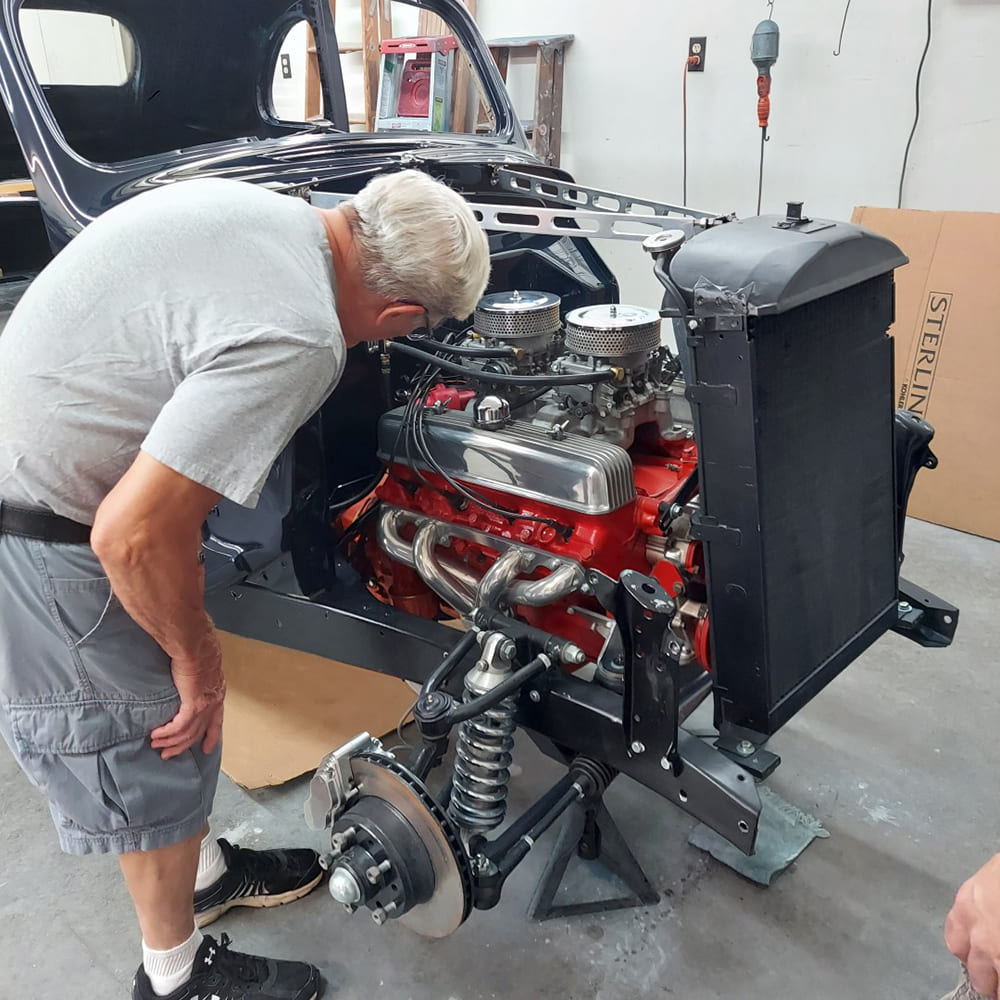 Mechanic leaning over the front clip of a 1936 Ford street rod inspecting the dual-quad carburetors, finned valve covers, radiator, and front coilover suspension.
