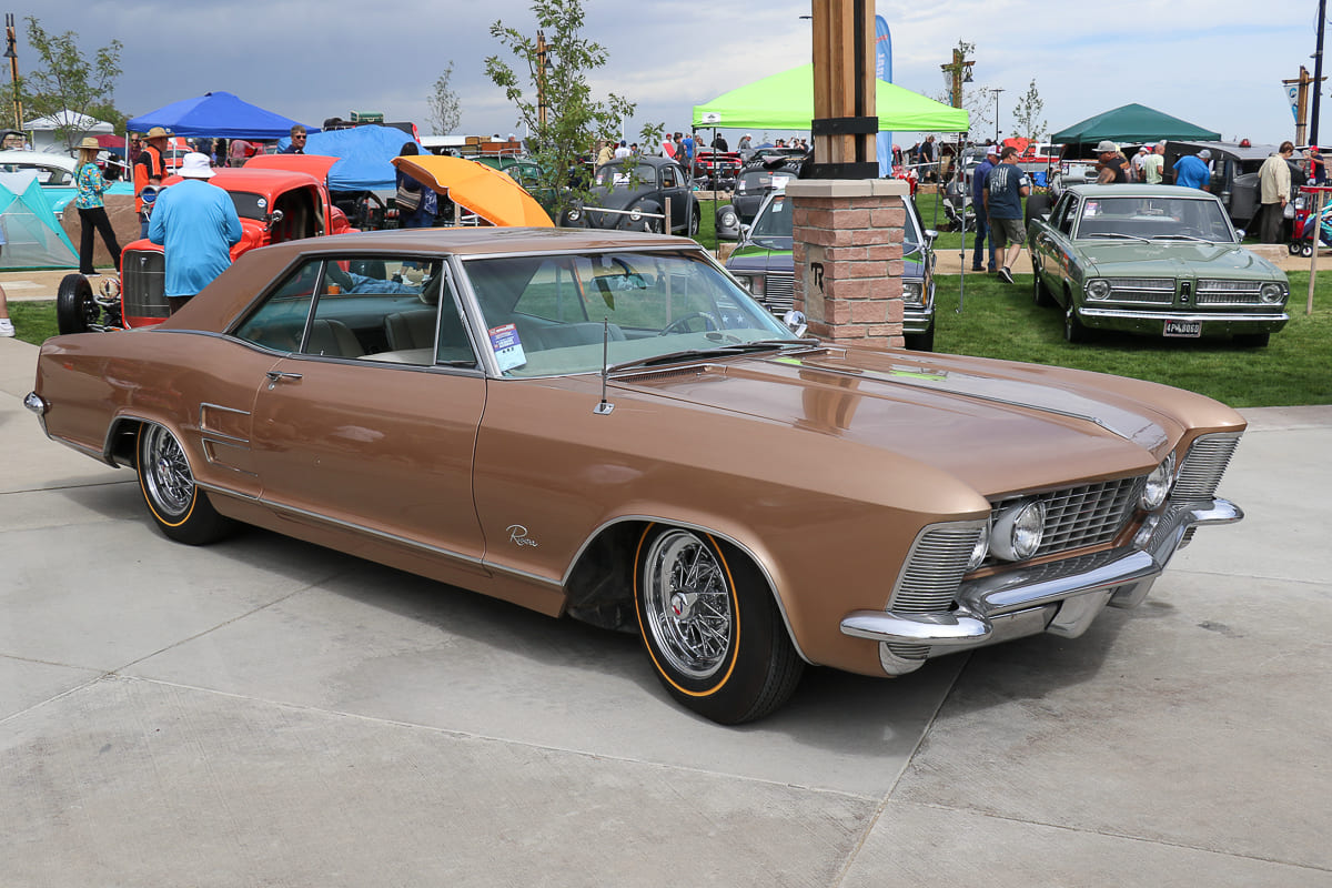 Front three-quarter angle of a lowered copper-gold 1964 Buick Riviera hardtop with wire wheels parked along an event concourse.