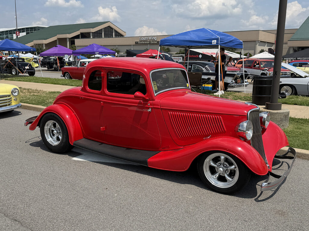Profile view of a vibrant red 1934 Ford five-window coupe street rod with louvered hood sides and polished chrome wheels in a car show parking row.