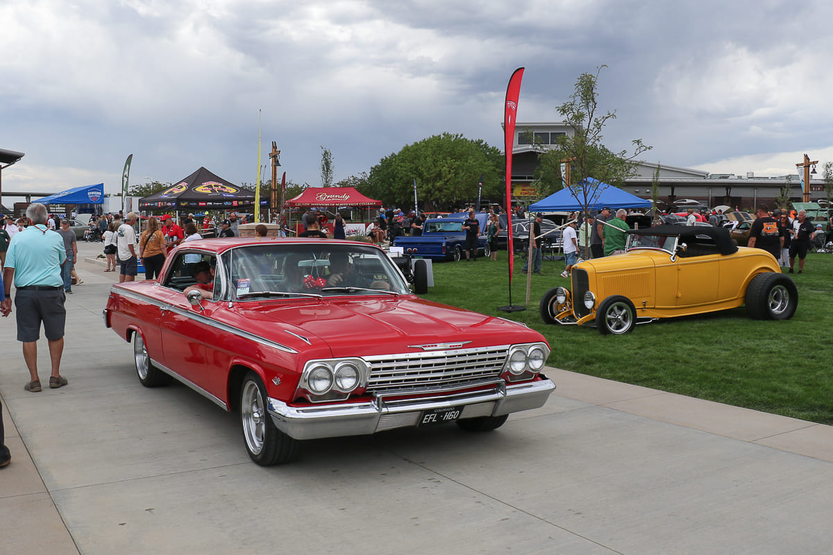 Front view of a bright red 1962 Chevrolet Impala hardtop driving on a show path past a parked yellow 1932 Ford roadster.