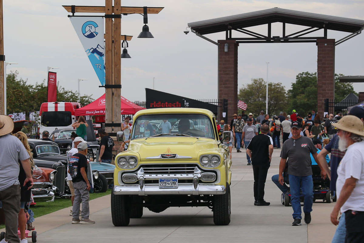 Head-on shot of a pale yellow 1958 Chevrolet Apache truck driving down a central walking plaza through the entrance gate structure.