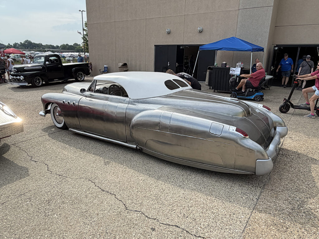 Rear three-quarter perspective of an ultra-low, chopped custom coupe in bare hand-formed steel featuring a white Carson-style top, wide whitewall tires, and fender skirts parked on concrete.