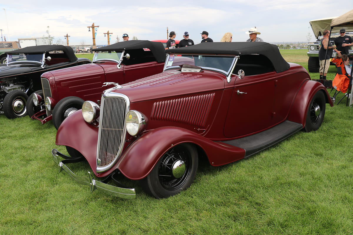 Front three-quarter view of a deep maroon 1934 Ford roadster with black soft top parked on grass beside a matching vintage coupe.