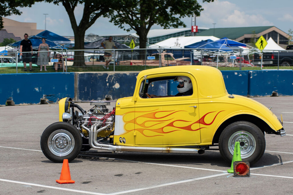 Side profile of a bright yellow 1932 Ford three-window coupe with traditional orange flames, open engine bay, and white headers navigating an autocross cone course.