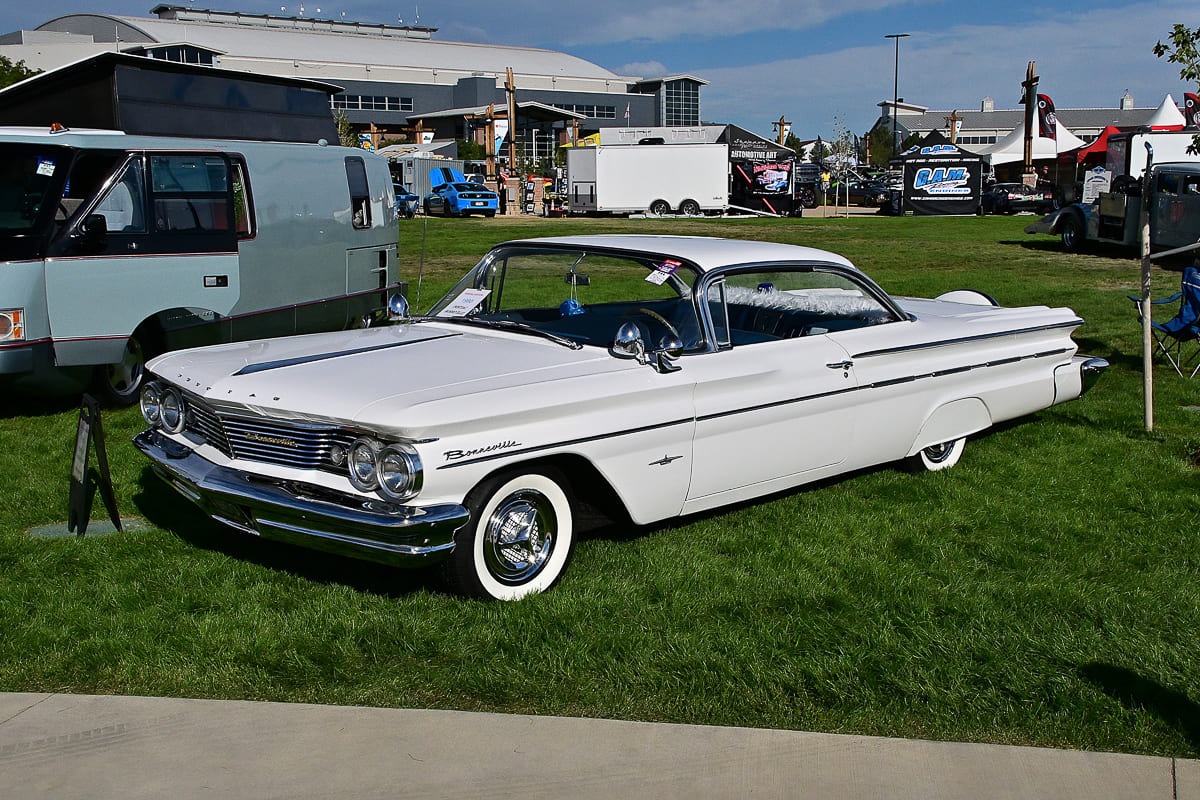 Front three-quarter view of an off-white 1960 Pontiac Bonneville sport coupe with wide whitewall tires parked on green grass at Good Guys Loveland Nationals.