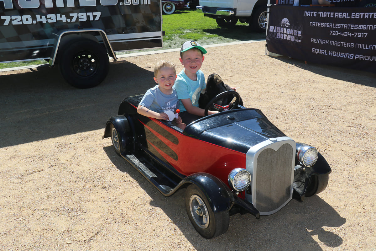 Two young smiling boys sitting inside a miniature black and red roadster pedal car parked on dirt beside an event trailer.