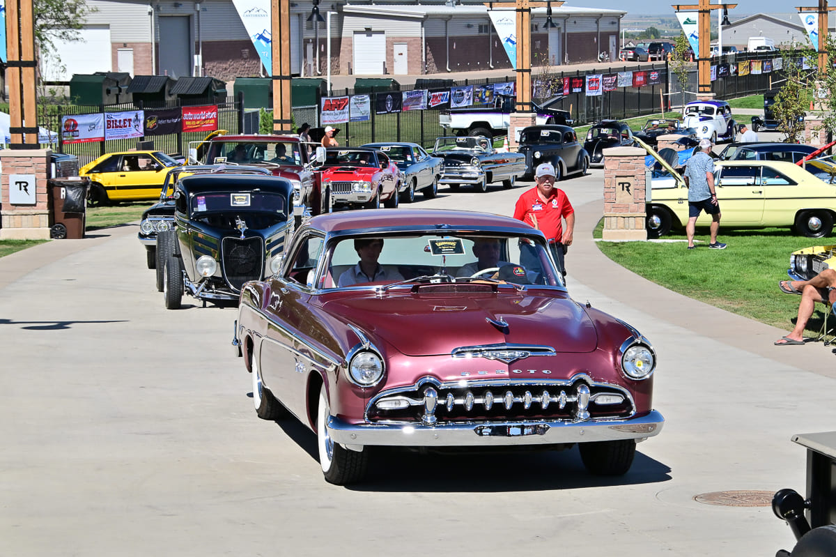 Front view of a burgundy 1954 DeSoto Firedome hardtop cruising down an event concourse leading a procession of vintage hot rods and customs.