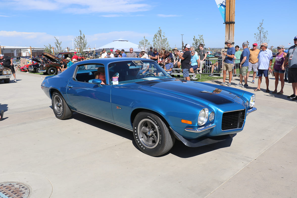 Front three-quarter view of a bright metallic blue 1970 1/2 Chevrolet Camaro RS/Z28 with black hood stripes cruising slowly past car show spectators