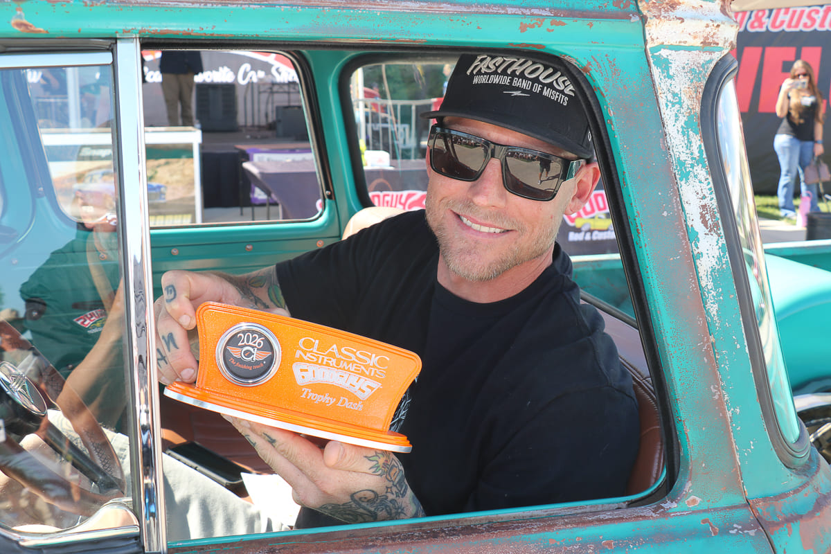 Driver in sunglasses and black hat smiling from the cab of his patina green vintage pickup while holding up a Classic Instruments Trophy Dash award plaque