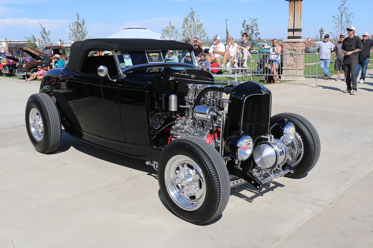Front three-quarter angle of a black 1932 Ford highboy roadster with polished chrome V8 engine, black soft top, and vintage wire wheels on concrete.