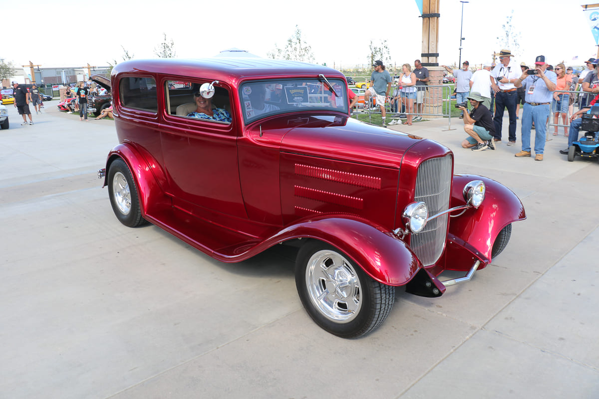 High front three-quarter view of a glossy candy apple red 1932 Ford Tudor sedan street rod rolling past spectators on an outdoor show concourse.