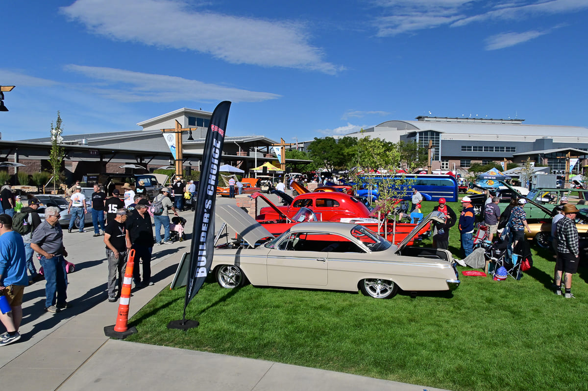 Outdoor grassy show field featuring custom classic cars and hot rods displayed around a black Builder's Choice flag and orange cones.