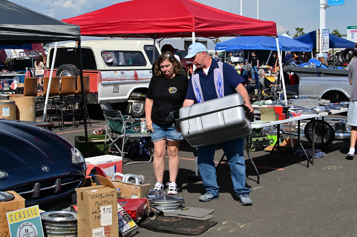 Couple browsing an outdoor automotive swap meet area with vintage hubcaps, exhaust mufflers, and a fuel tank under a red canopy.