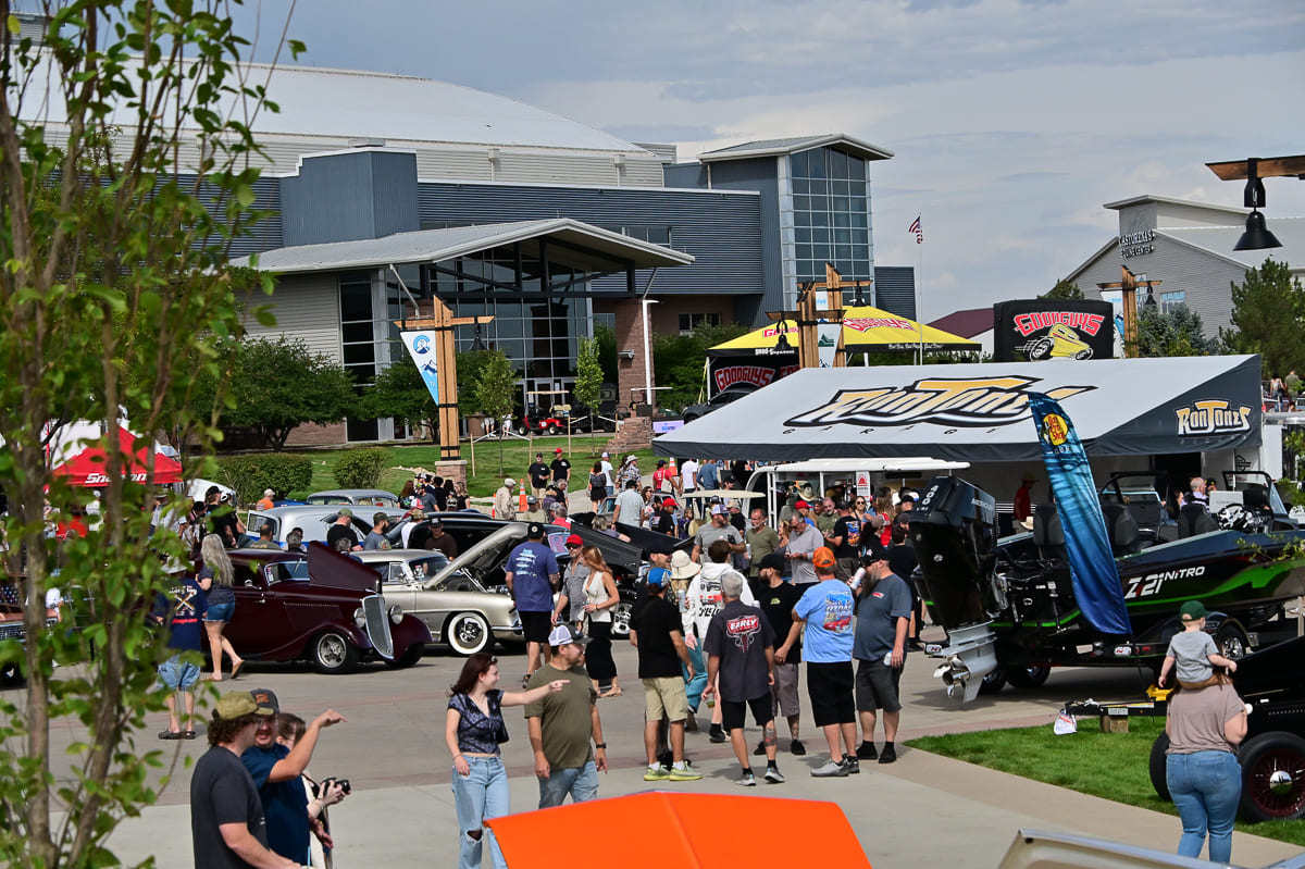 Elevated view of crowds walking the outdoor vendor midway between custom boats, trailers, and classic hot rods parked outside an event arena.