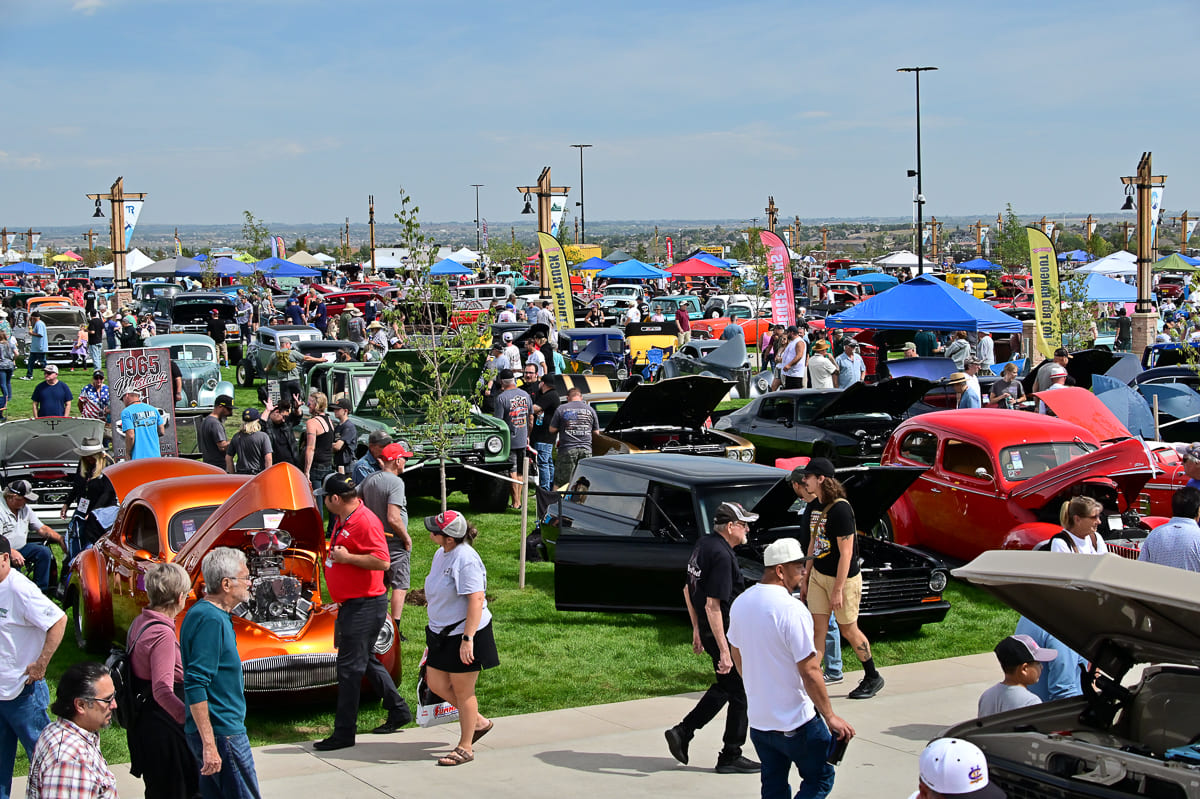 Wide elevated panoramic perspective of hundreds of classic cars and hot rods displayed across a sprawling grassy lawn under a sunny sky.