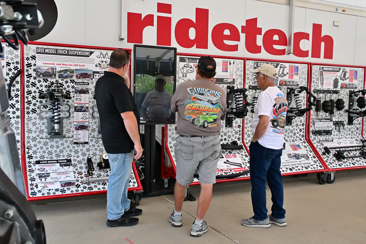 Three attendees examining suspension components and digital displays inside an indoor Ridetech booth at a car show.