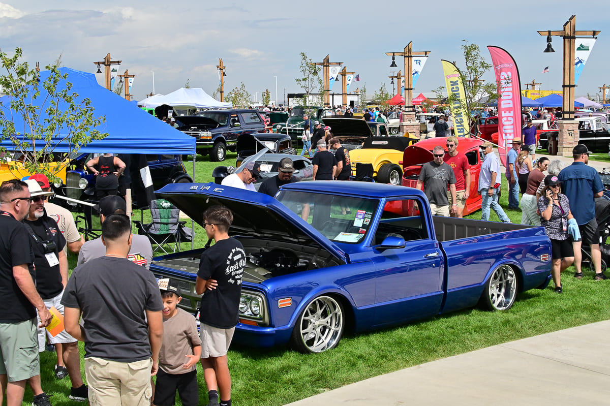 Front three-quarter view of a bright metallic blue classic GMC pickup truck with an open hood and chrome wheels surrounded by car enthusiasts.