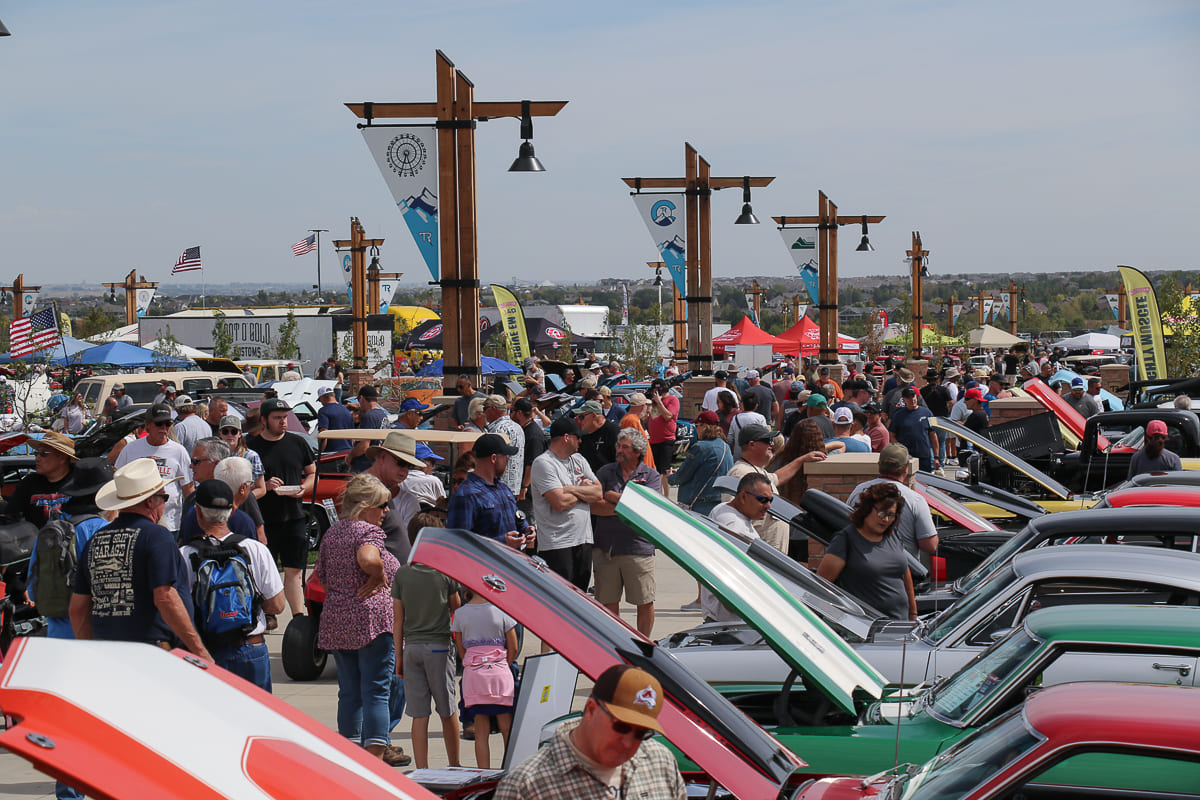 Busy pedestrian concourse lined with open-hood classic muscle cars and spectators walking beneath tall wooden banner pillars.