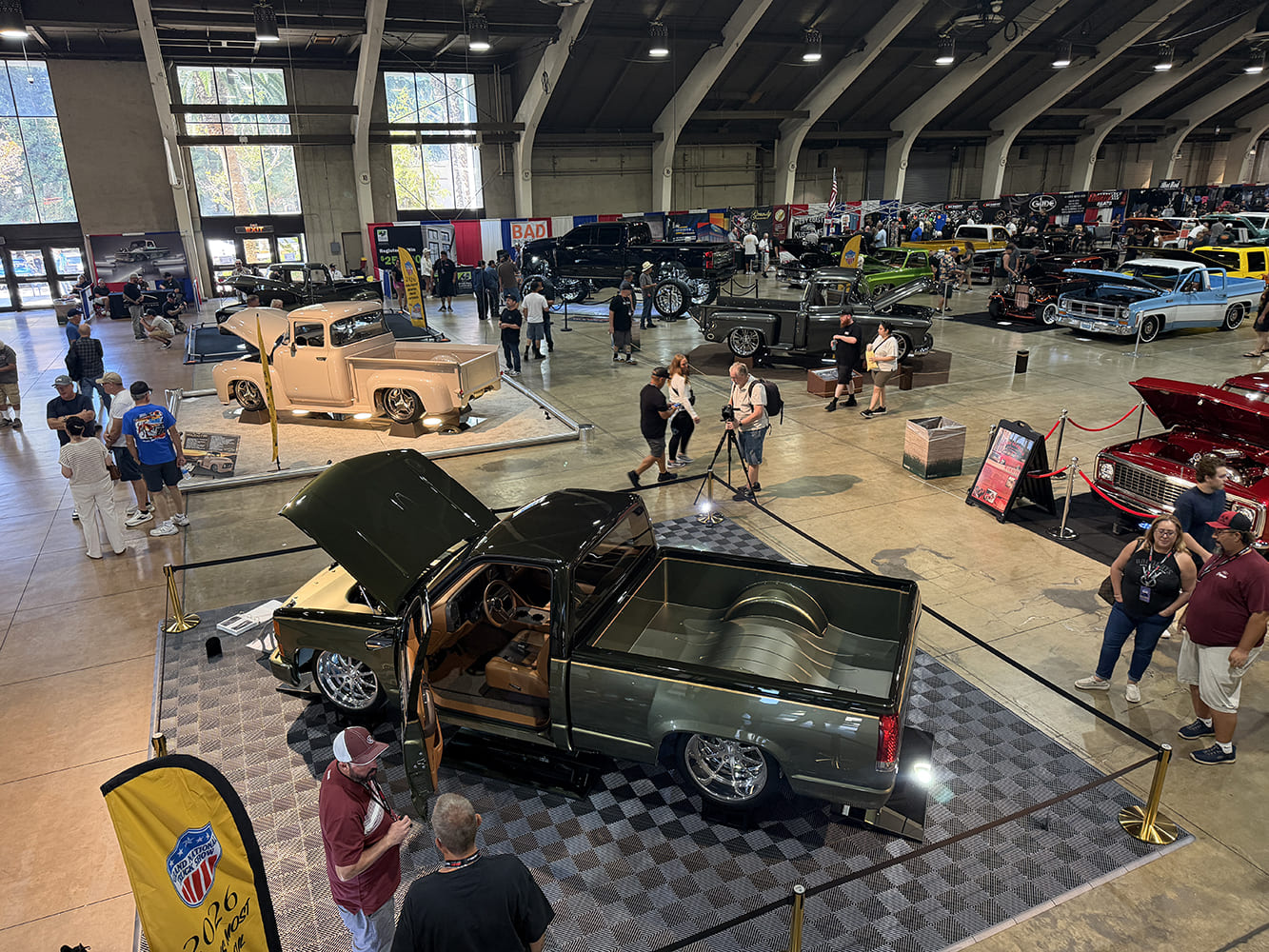 High-angle overview of Expo Hall at the Grand National Truck Show showing custom lowered and lifted trucks, vendor displays, and attendees inside the venue.