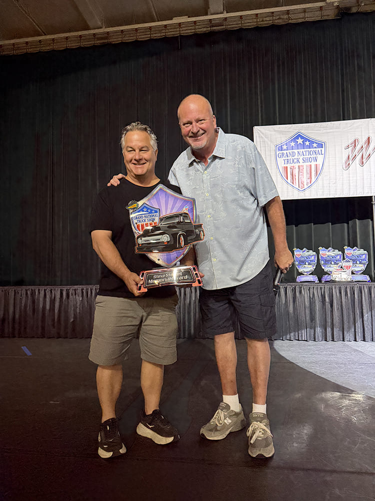 Stage presenter smiling alongside the award recipient holding the custom Steve Stillwell Memorial Award shield at the Grand National Truck Show ceremony.