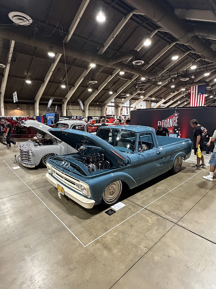 Front three-quarter angle of a lowered pastel blue Ford F-100 pickup truck with its hood open to showcase a multi-carburetor engine on the show floor.