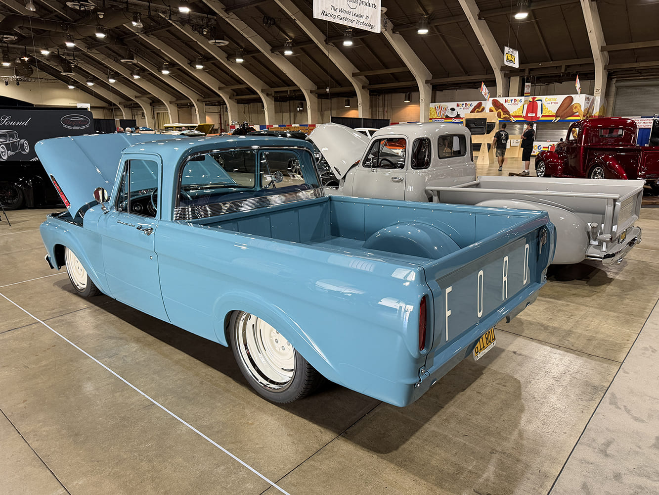 Rear three-quarter perspective of a lowered light blue classic Ford pickup showing white steel wheels, smooth tailgate with white lettering, and raised wheel tubs.