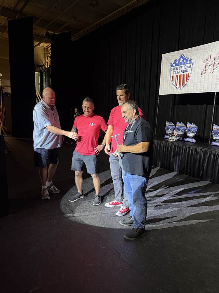 Event organizers and builders smiling on stage during an awards presentation in front of the Grand National Truck Show backdrop.