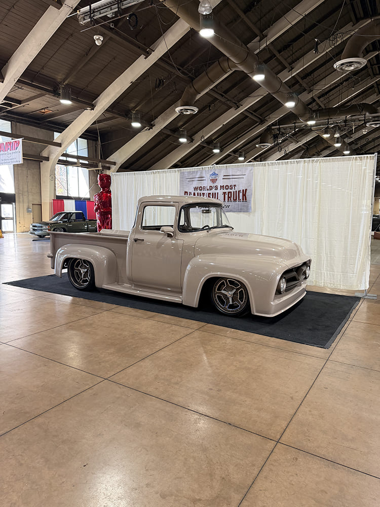 Front three-quarter angle of a slammed light gray Ford F-100 custom pickup sitting on carpet in front of the World’s Most Beautiful Truck backdrop.