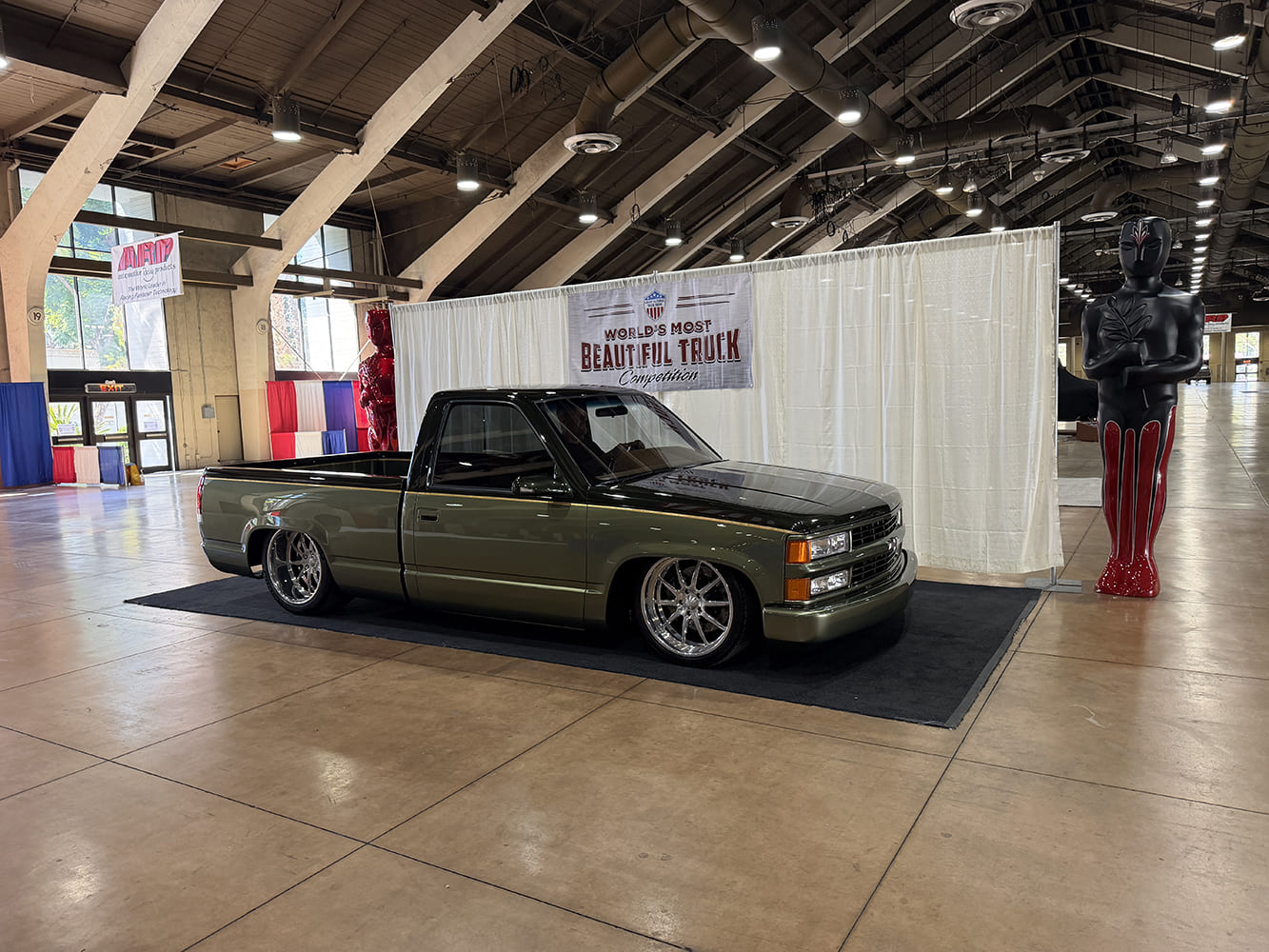 Driver-side profile of a lowered two-tone olive green and black Chevrolet C1500 OBS pickup on polished wheels in front of event stage drapes.