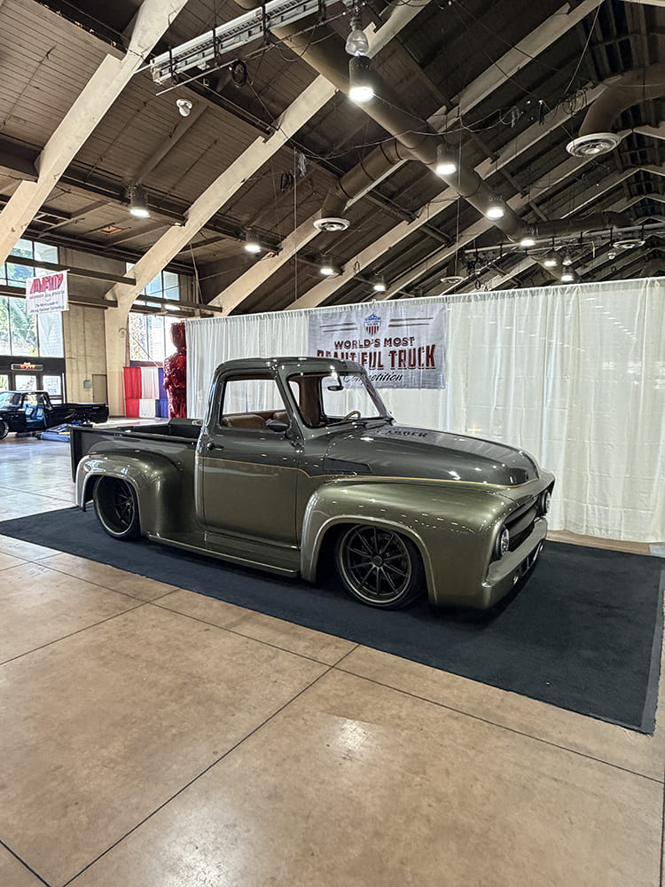 Front three-quarter view of a slammed metallic olive-bronze Ford F-100 stepside pickup on modern gray performance wheels in an exhibition hall.
