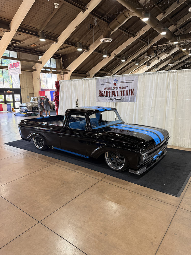 front three-quarter view of a lowered gloss black Ford F-100 unibody truck with dual electric blue racing stripes and dark multi-spoke wheels.
