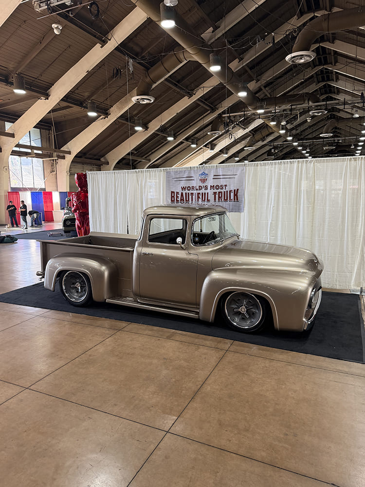 Profile view of a slammed champagne metallic 1956 Ford F-100 big-window pickup resting on black carpet in front of the competition stage banner.
