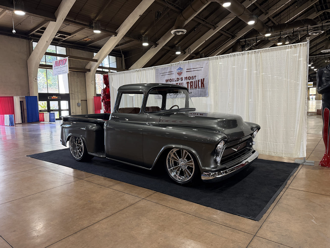 front three-quarter view of a slammed metallic dark charcoal 1957 Chevrolet pickup with deep chrome billet wheels displayed inside the expo building.