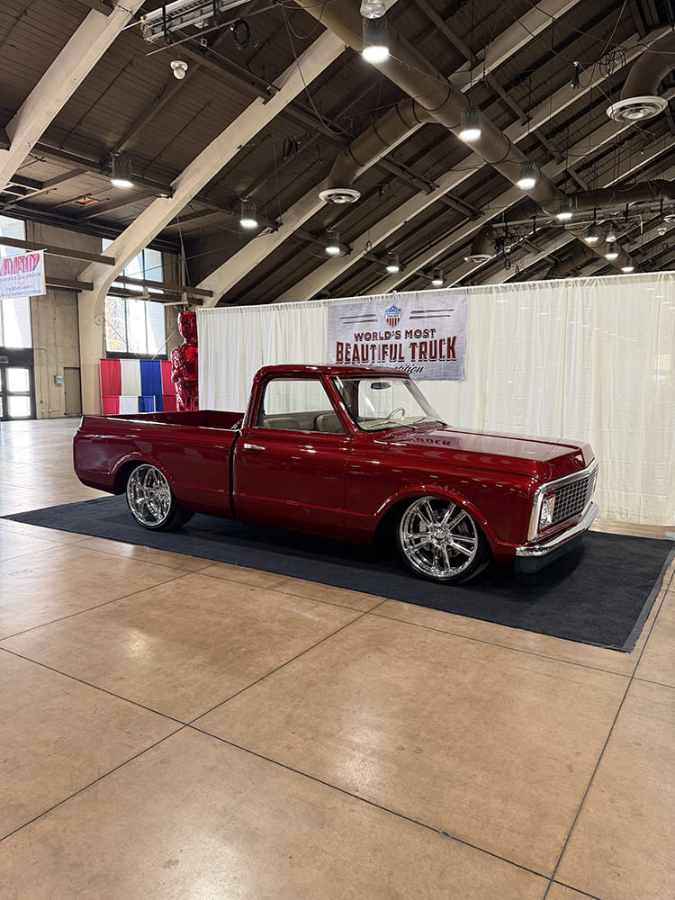 Side three-quarter perspective of a slammed deep candy red Chevrolet C10 pickup truck on large chrome wheels parked on black display carpet.