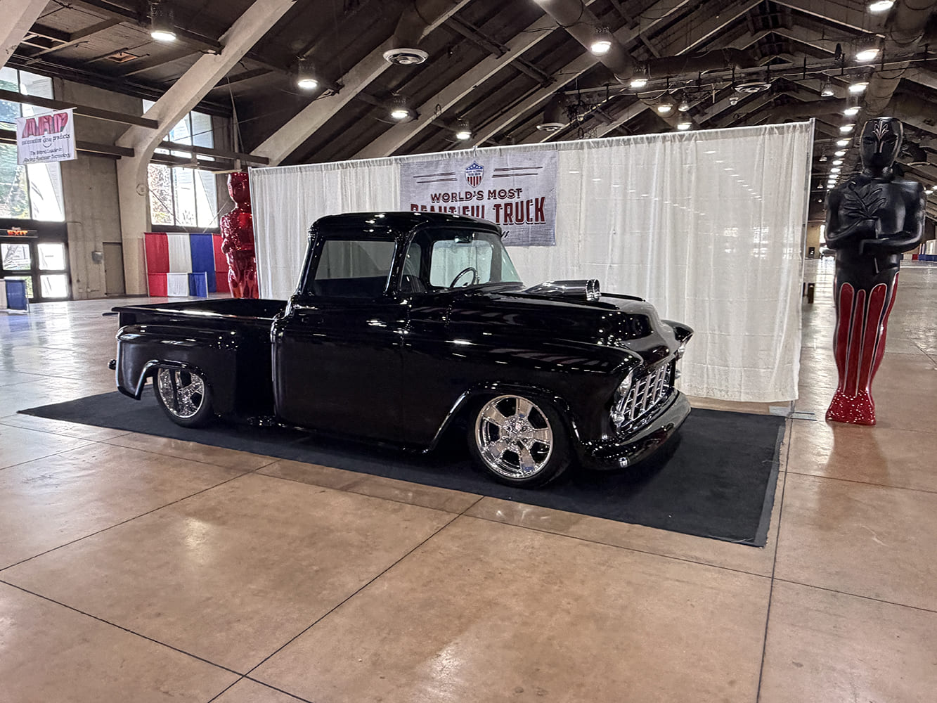 Low-angle profile of a jet-black 1955 Chevrolet pickup featuring a hood cowl intake scoop and deep chrome wheels in front of the World's Most Beautiful Truck backdrop.